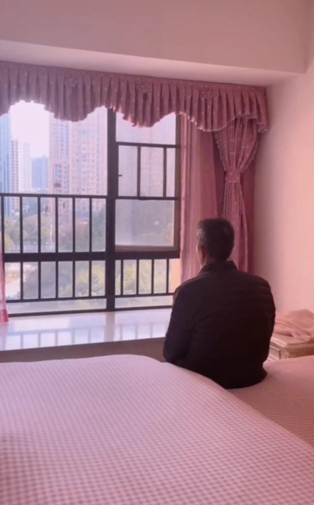 Elderly Asian man drinking tea and looking out of a bedroom window at a public square